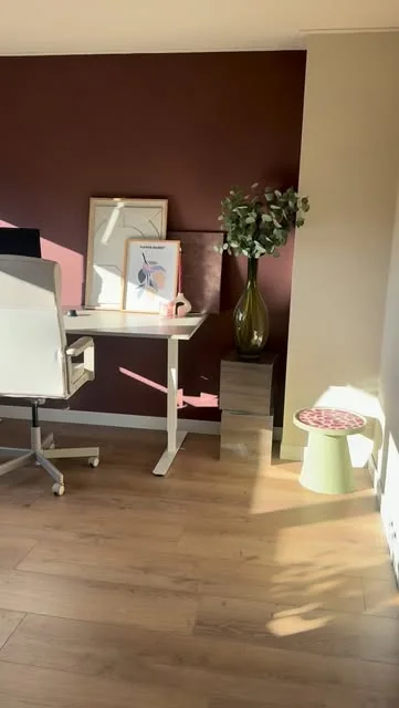 Werkplek met een witte bureaustoel voor een donkerrode wand met kunst en een plant