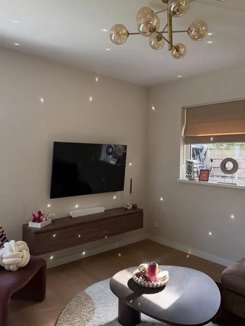 Woonkamer met een grijze hoekbank, roze kussens, een ronde donkere salontafel en een wolkvormig vloerkleed